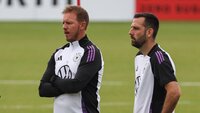 Drei Fußballtrainer stehen auf einem Trainingsplatz in offizieller Teamkleidung.