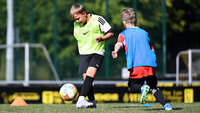 Kind dribbelt beim E‑Jugend‑Fußballtraining, weiteres Kind läuft mit; unscharfe Tore im Hintergrund.