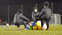 Kinder im Wintertraining auf dem Fußballplatz sammeln bunte Bälle im Kreis.