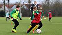 Mehrere Kinder spielen Fußball auf einem Trainingsplatz; ein Kind im roten Leibchen dribbelt den Ball, zwei Kinder im grünen Leibchen laufen mit, im Hintergrund beobachtet eine erwachsene Person.