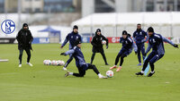 Mehrere Schalke-04-Spieler trainieren bei kühlem Wetter, während ein Spieler im Vordergrund zum Ball grätscht.