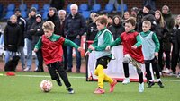 Kinder spielen Fußball auf einem Platz, Zuschauer stehen am Spielfeldrand.