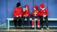 Kinder in roten Trainingsjacken sitzen auf einer Bank und halten Trinkflaschen in den Händen.