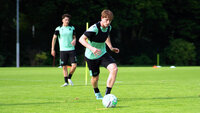 Ein Fußballspieler im weißen und schwarzen Trainingsoutfit dribbelt während einer Trainingseinheit auf einem Rasenplatz mit Übungshilfen im Hintergrund.