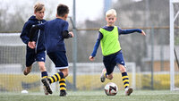 Drei Kinder spielen Fußball auf einem Kunstrasenplatz; ein Kind im neongrünen Leibchen dribbelt den Ball, während zwei Kinder in dunkelblauen Trikots heraneilen.